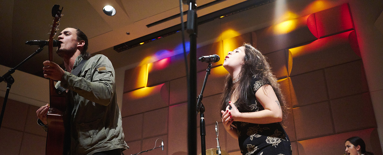 Vocalists sing into microphones on stage while a man plays the guitar