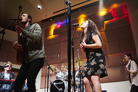 Vocalists sing into microphones on stage while a man plays the guitar